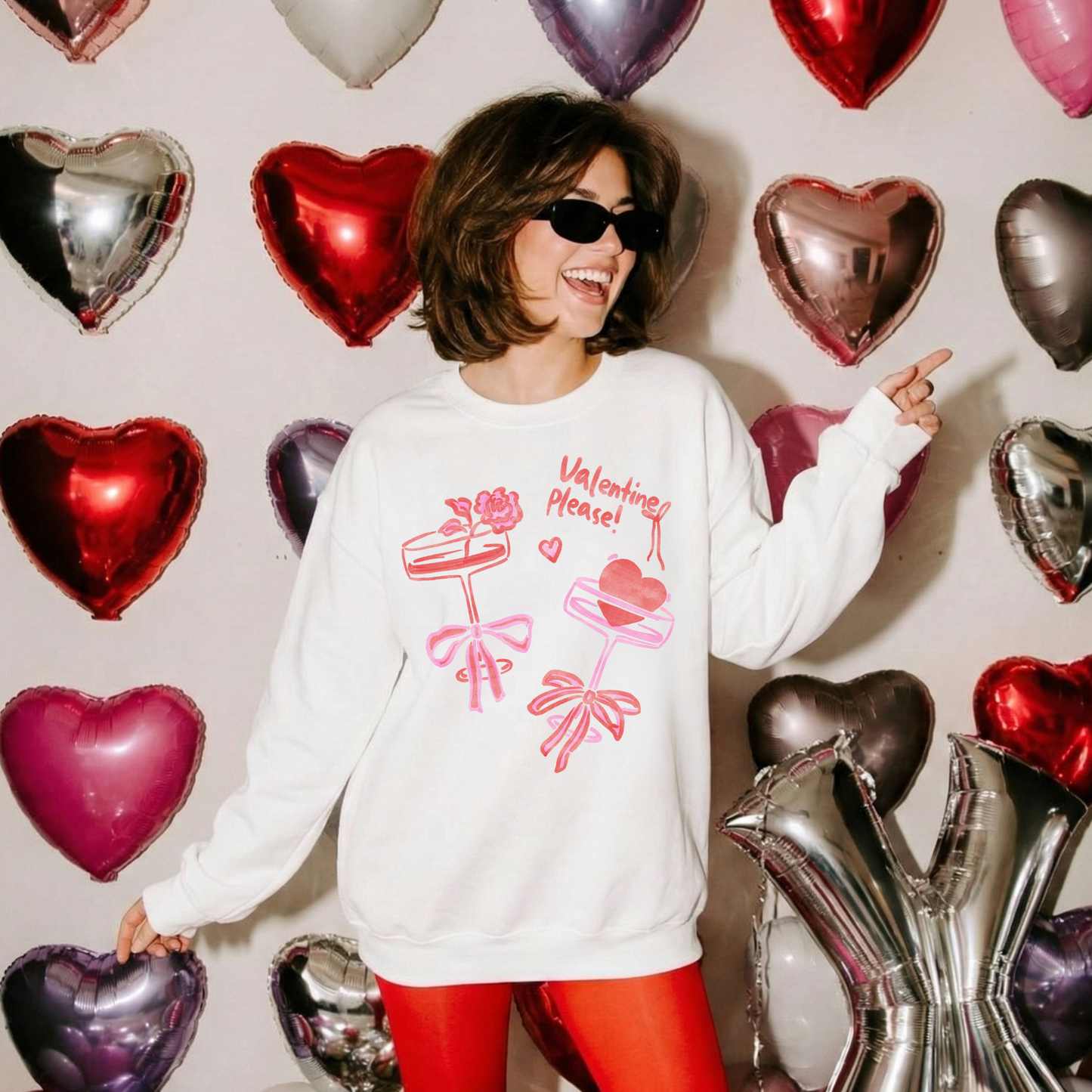 Woman wearing a white Valentine’s crewneck with pink and red bow designs, standing in front of red and pink heart balloons and smiling.