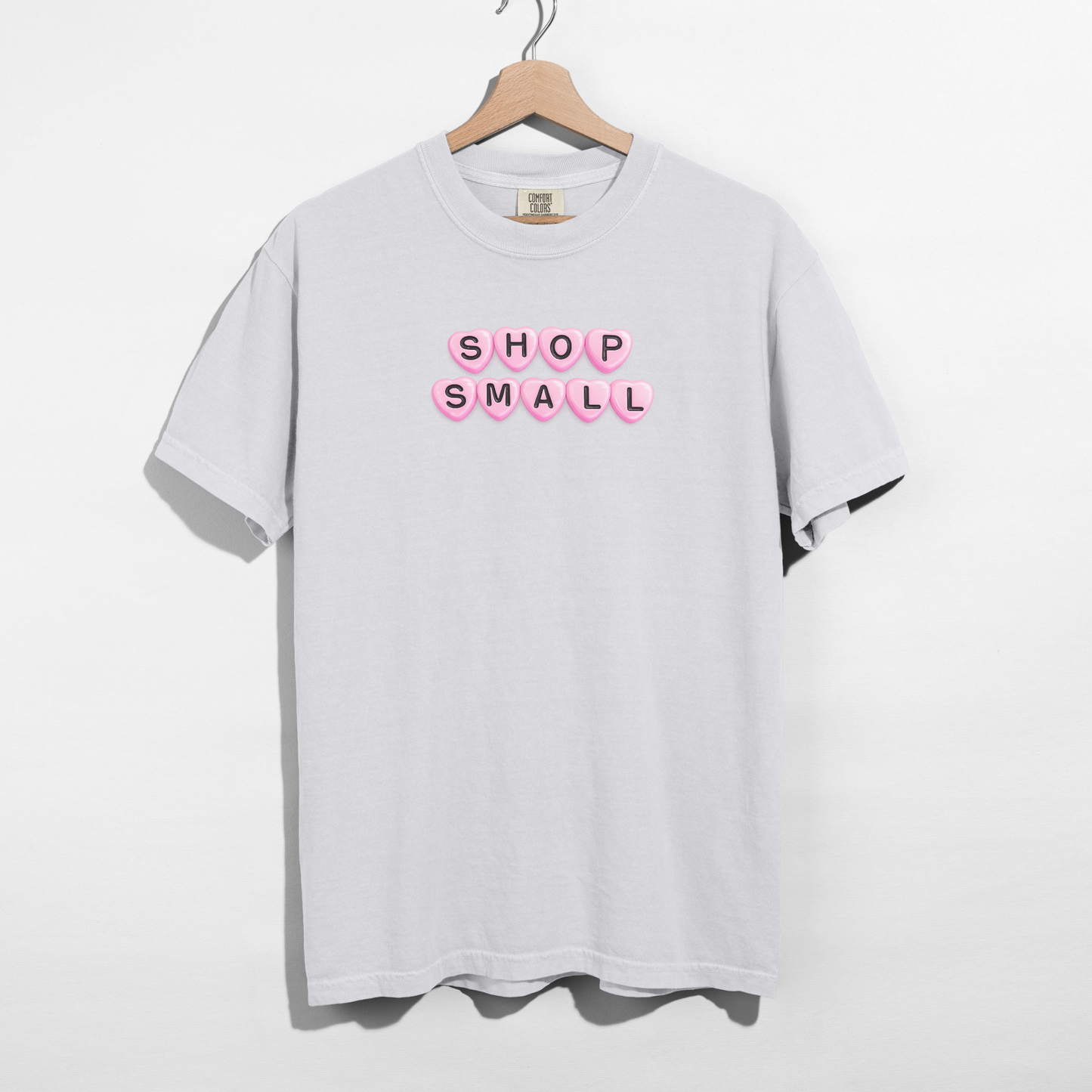 White t-shirt with 'SHOP SMALL' text in pink on a hanger against a white background