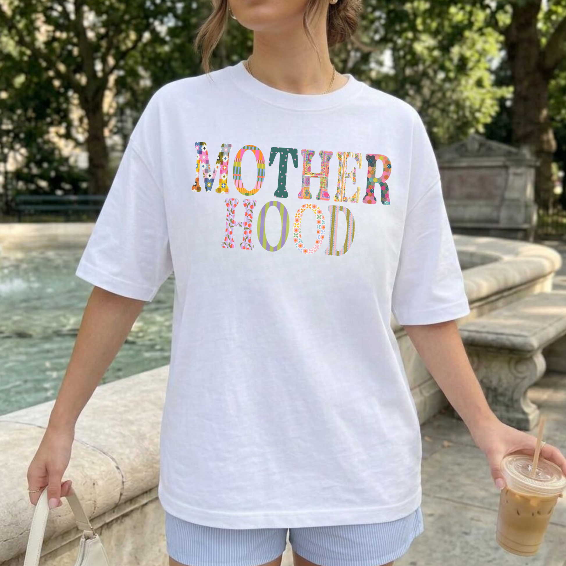 Woman wearing oversized white motherhood graphic tee with colorful patchwork lettering near outdoor fountain while holding iced coffee.