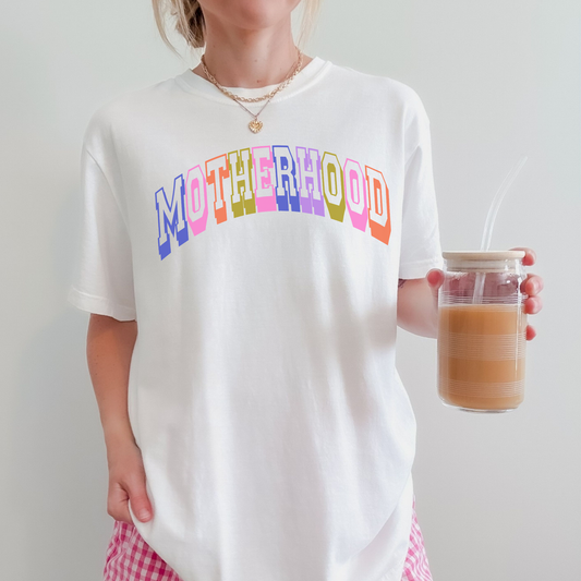 Woman wearing a white Comfort Colors 1717 graphic tee featuring colorful retro collegiate “Motherhood” lettering while holding iced coffee.