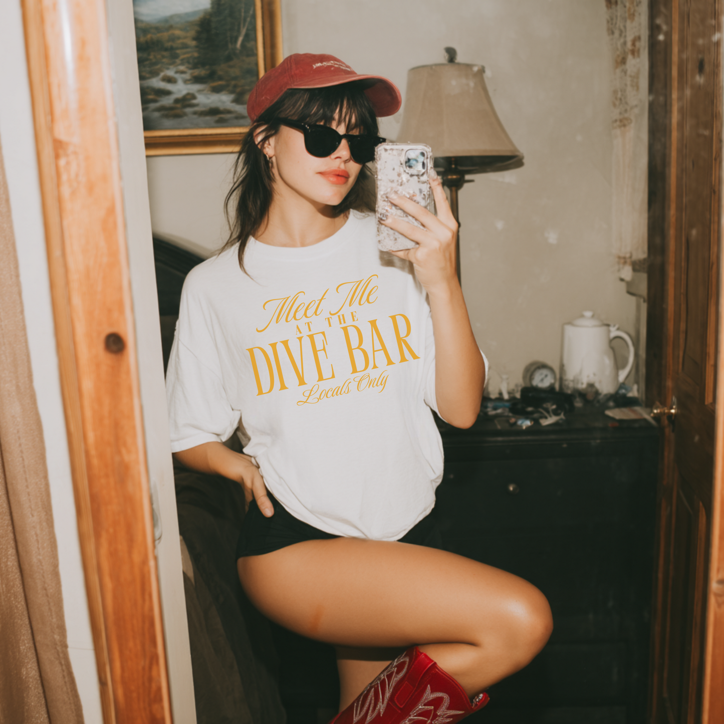Woman taking a mirror selfie wearing a white t-shirt with yellow text and a red cap.