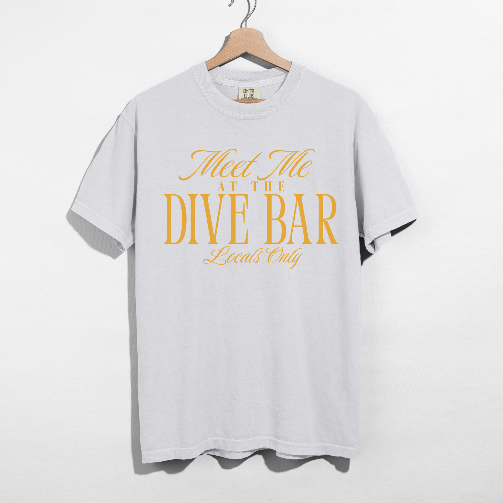 White t-shirt with yellow text 'Meet Me at the Dive Bar Locals Only' on a hanger against a white background