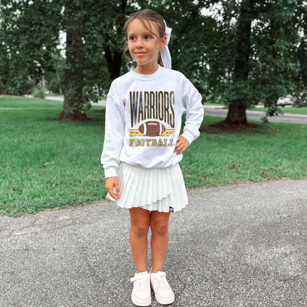 Young girl wearing a 'Warriors Football' sweatshirt and white skirt standing on a path with greenery in the background.