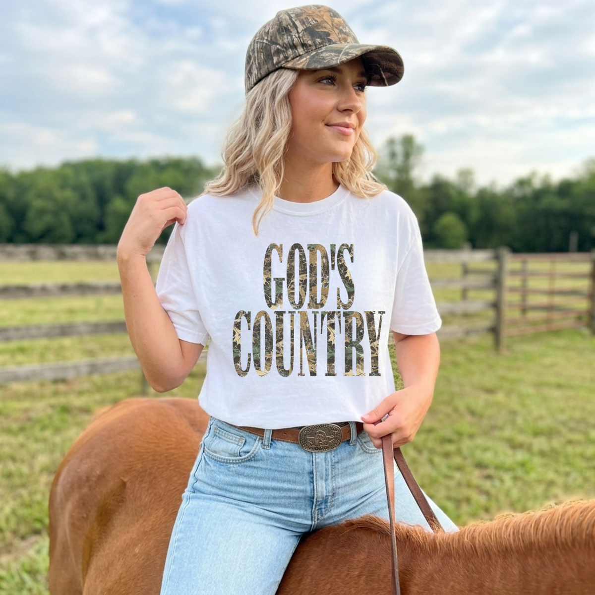 God’s Country camo graphic tee in white styled with light wash jeans and camouflage hat on horseback
