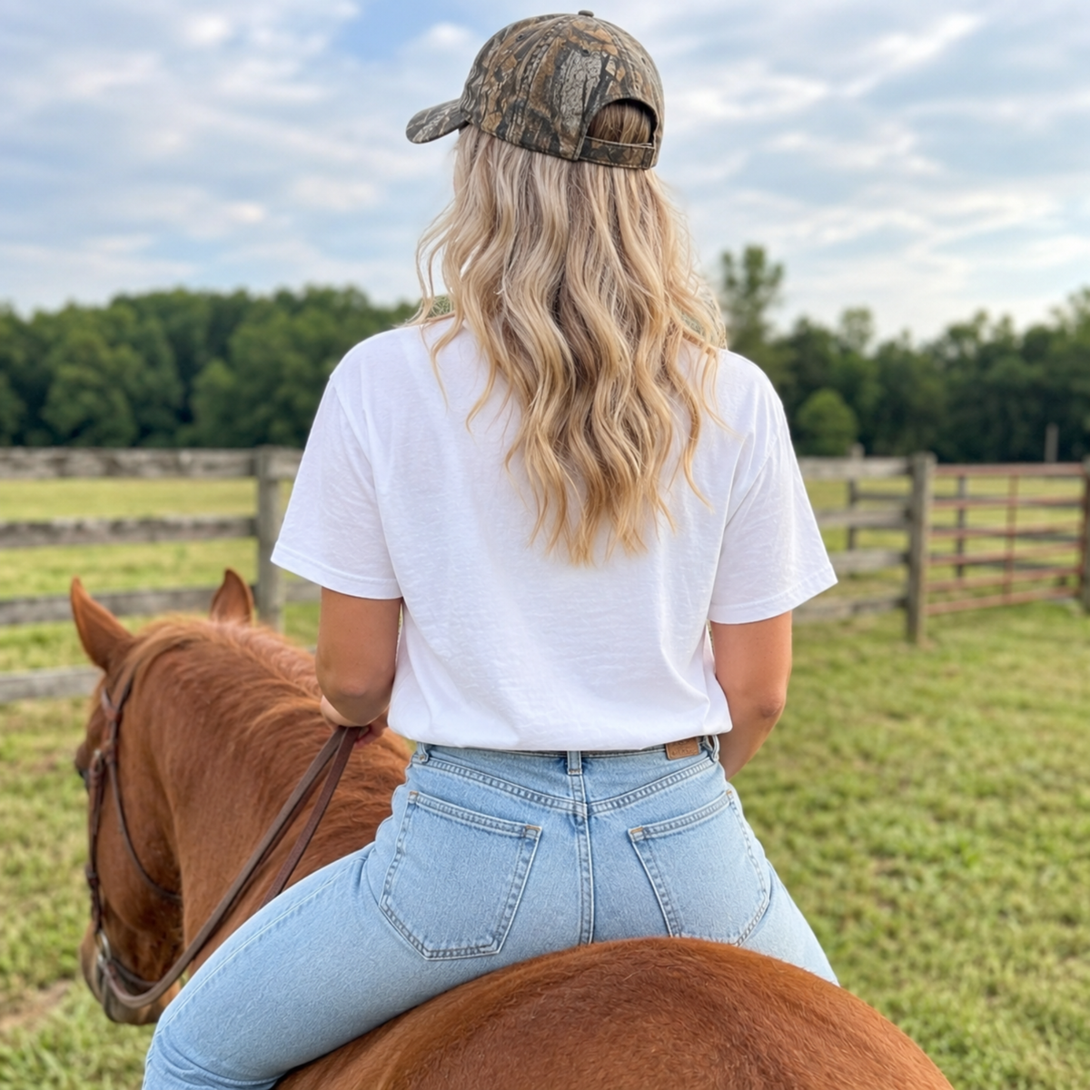 Back view of white God’s Country graphic tee styled with jeans and camouflage hat while riding a horse