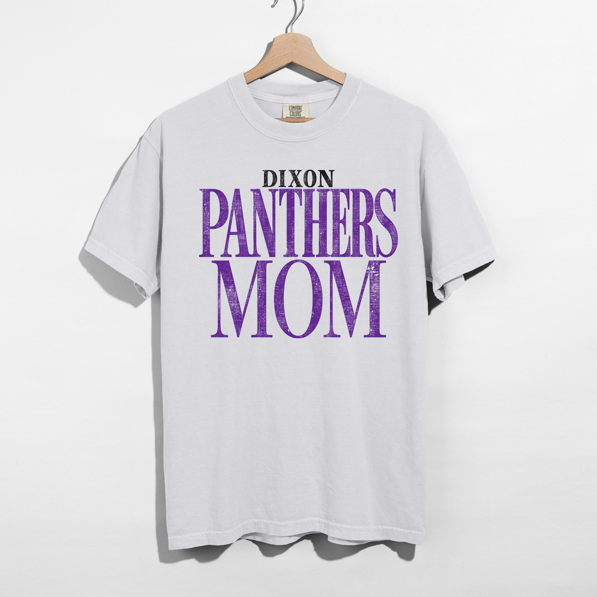 A white short-sleeve t-shirt hanging on a wooden hanger against a white wall. The shirt features a graphic print on the front that reads 'DIXON PANTHERS MOM' in black and purple distressed lettering.