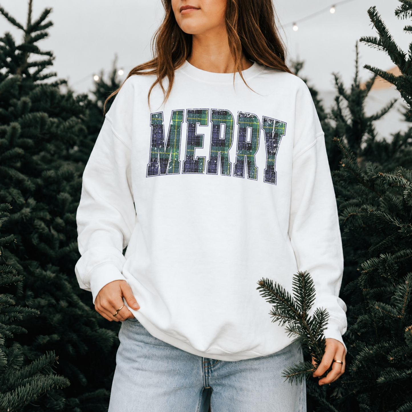 Woman wearing a white Lane Seven crewneck sweatshirt with vintage distressed navy and green tartan plaid Merry graphic at a Christmas tree farm.