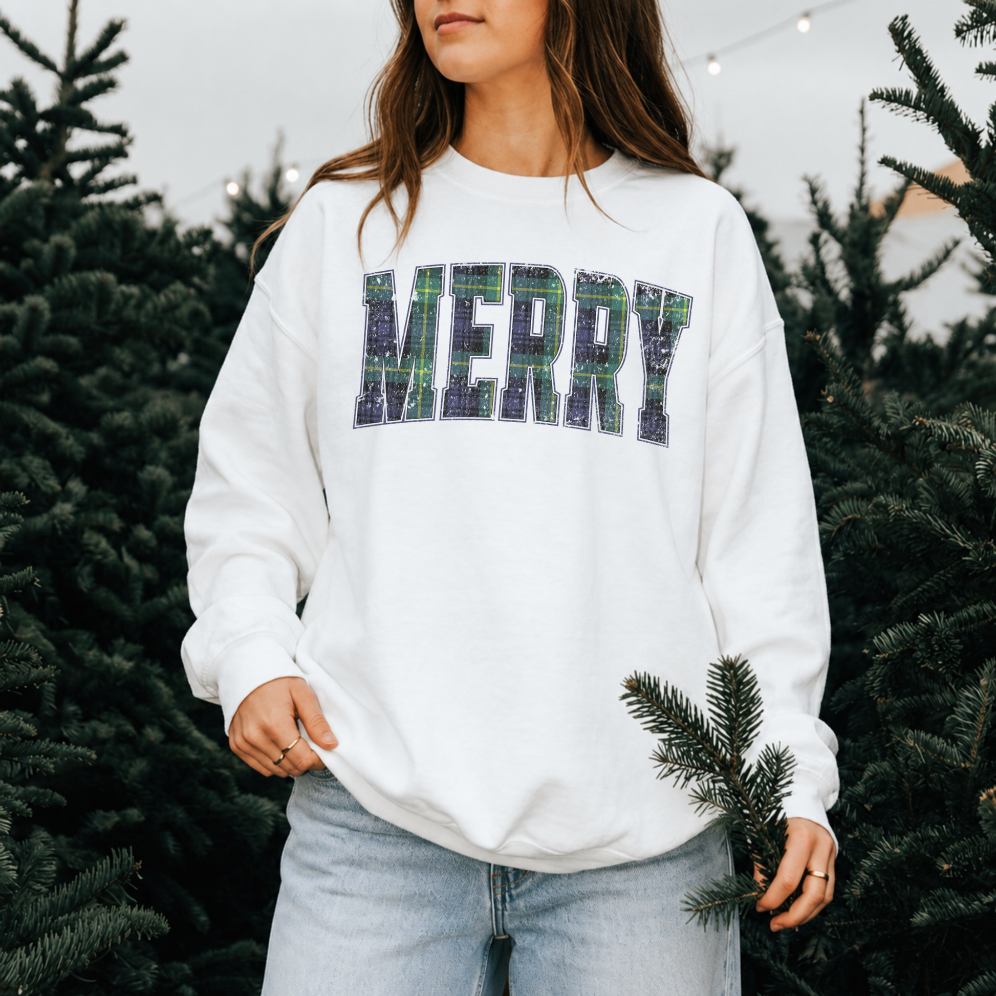 Woman wearing a white Lane Seven crewneck sweatshirt with vintage distressed navy and green tartan plaid Merry graphic at a Christmas tree farm.