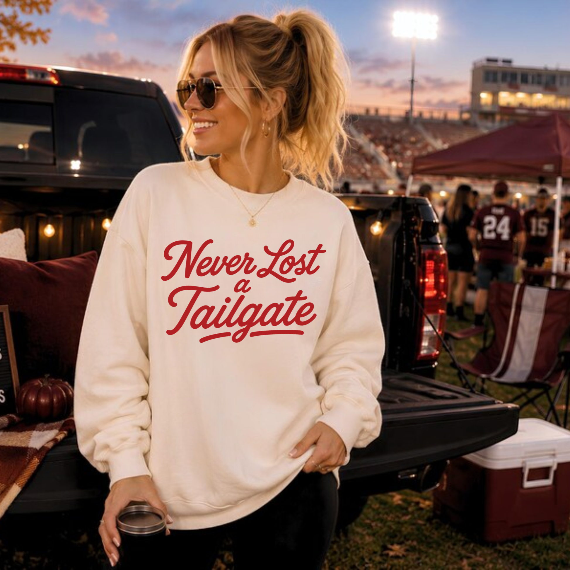 Woman wearing a white "Never Lost a Tailgate" football sweatshirt while tailgating before a football game at sunset with trucks and stadium lights in the background.