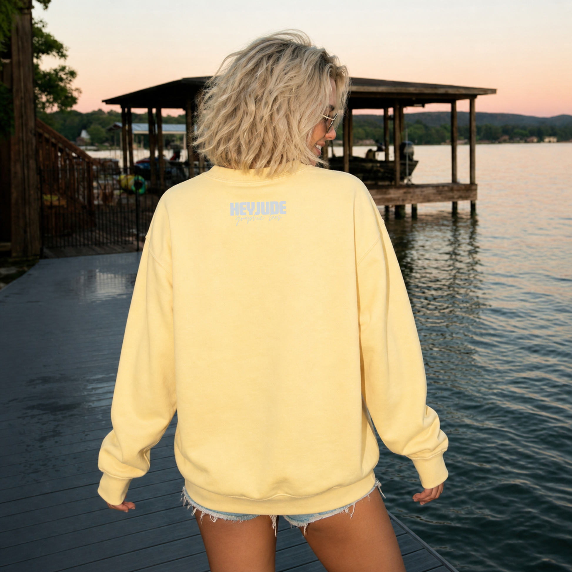 Person wearing a yellow sweatshirt with text on the back, standing by a lake with a dock.